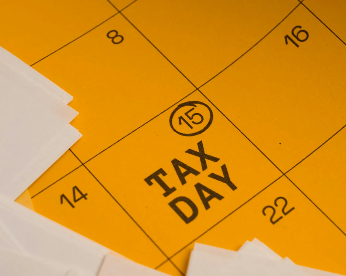 Close-up of a yellow business calendar with the 15th circled and marked 'TAX DAY,' white tax forms stacked alongside.
